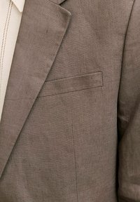 Közeli kép barna lenvászon blézerről, melyen beszabott gallér látható, krémszínű kötött felső fölött, mellzsebbel és anyagtextúrával.