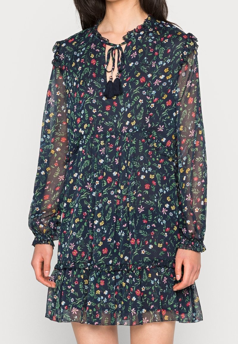 Robe florale bleu marine avec manches longues transparentes, poignets élastiques, ourlet à volants et col à nouer avec des pompons.