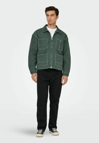 Chaqueta de denim verde con dos bolsillos frontales tipo parche, costuras blancas y cierre de cremallera, combinada con pantalones negros y zapatillas.