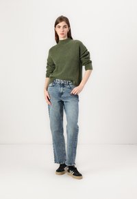 Groene gebreide trui, comfortabele lichtblauwe jeans en zwarte sneakers met tan kleurige zolen. Model staat tegen een neutrale achtergrond.