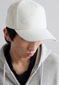 Hvid baseballcap lavet af tekstureret stof, med et forhøjet logo. Buet skygge, struktureret form. Båret lidt skråt fremad.