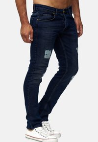 Slank blå denimjeans med slidte lapper, med et klassisk fem-lomme design og gule hardware-detaljer, parret med hvide sneakers.