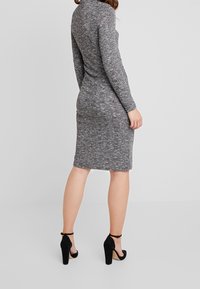 Polvipituinen, vartalonmyötäinen harmaa mekko, jossa on teksturoidun kuvio, pitkät hihat ja korkea kaulus, yhdistetty mustiin korkokenkiin.