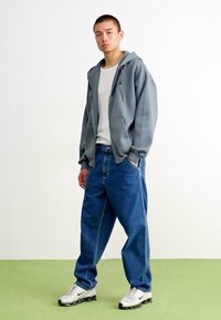 Gray hooded sweatshirt, white t-shirt, loose blue jeans, and white athletic shoes with black accents, standing against a plain background.