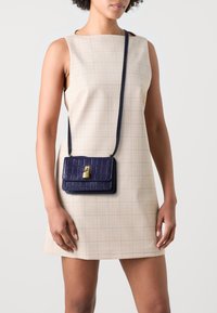 Borsa in pelle stampata coccodrillo blu navy con chiusura dorata, dotata di una lunga tracolla. Indossata con un vestito a quadri beige chiaro, che mette in risalto un design minimalista.