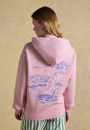 Kvinde med blondt hår har en lyserød hættetrøje med blåt sejlbåd- og kystlandsbyskilt med teksten "Great Vibes Joules," kombineret med grønne striped bukser.