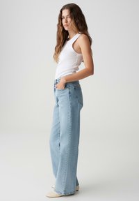 Light blue flared jeans made of denim, featuring a high waist, front pockets, and a slightly relaxed fit. Paired with a fitted white tank top.