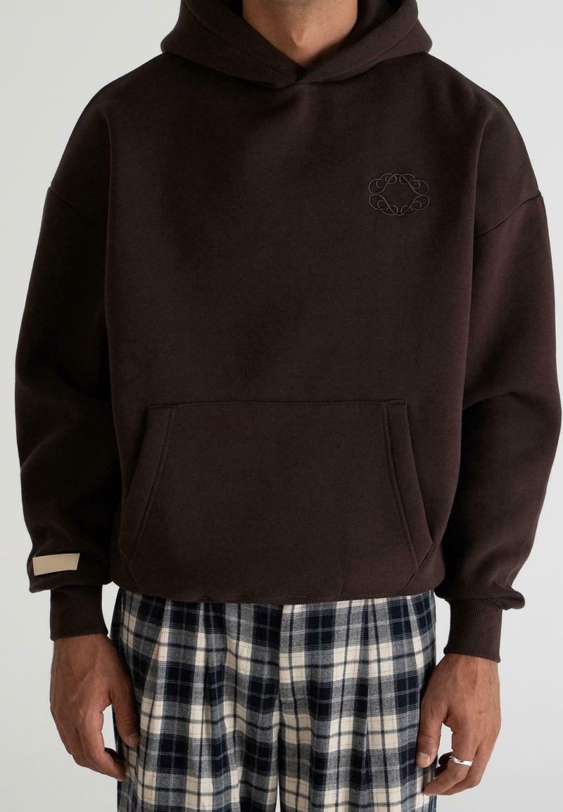 Homme portant un sweat à capuche marron foncé avec poche avant et petit logo brodé, associé à un pantalon à carreaux noir et blanc.