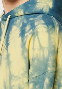 Gros plan sur une personne portant un sweat à capuche tie-dye bleu et jaune avec des cordons blancs sur la capuche.