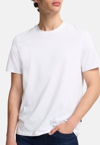 Hvid bomulds-t-shirt med rund hals og korte ærmer, som har en let stretch. Bruges sammen med mørkeblå jeans, hænderne i lommerne.