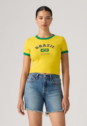 Kvinde iført en tætsiddende gul crop top med grøn kant og teksten "Brasil" samt flag, kombineret med højtaljede denimshorts med opsmøgede kanter.