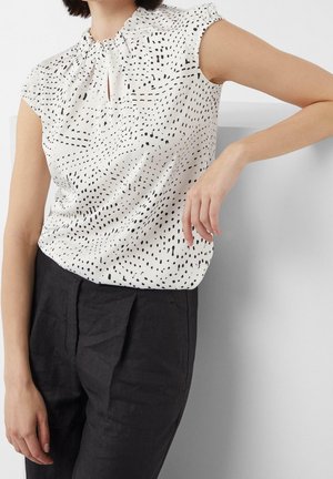 Frau trägt eine ärmellose weiße Bluse mit schwarzen abstrakten Punkten und hoch taillierte schwarze plissierte Hose, lehnt sich an eine weiße Oberfläche.