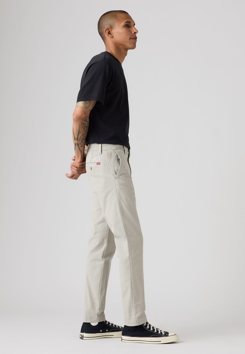 Homme avec des tatouages portant un t-shirt noir, un pantalon beige et des baskets noires, debout de profil, les mains jointes dans le dos.