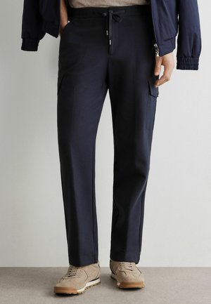 Mann trägt marineblaue Cargohose mit Kordelzug in der Taille und beige Wildledersneaker, steht vor einer schlichten hellen Wand.