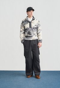 Noor mees, kes kannab musta mütsi, mustri ja musta lõuendiga fliisist pluusi, laiu musti töösäärekangas pükse ja pruune kingi, seisab sinise vaiba peal valge seina taustal.