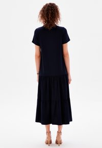 Person med lockigt hår som bär en kortärmad svart, lager-på-lager, midiklänning och hudfärgade högklackade sandaler, stående vänd bort mot vit bakgrund.