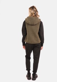 Chaleco de forro polar verde oliva con capucha, combinado con una camiseta negra de manga larga y pantalones jogger negros. Presenta una textura suave y un ajuste relajado.