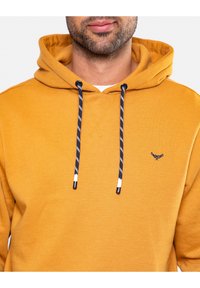 Felpa color senape con tasca frontale a canguro, cordini a righe bianche e nere, e un piccolo accento ricamato. Tessuto in misto cotone.