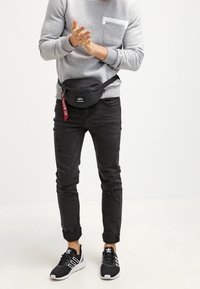 Svart midjeväska med dragkedja, bärs över en grå sweatshirt med bröstficka, matchad med svarta slitna jeans och svarta sneakers.