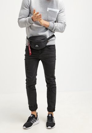 Riñonera negra con cremallera, llevada sobre un sudadera gris con un bolsillo en el pecho, combinada con jeans negros rasgados y zapatillas negras.