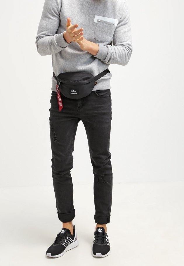 Svart midjeväska med dragkedja, bärs över en grå sweatshirt med bröstficka, matchad med svarta slitna jeans och svarta sneakers.