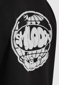 Camiseta de algodón negra con un gran gráfico blanco de una calavera con manos y la palabra "SMILODON" en una fuente estilizada.