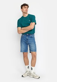 Teal katoenen T-shirt met klein logo, gecombineerd met blauwe denim shorts en lichtgrijze sneakers. Model staat met gekruiste armen tegen een witte achtergrond.