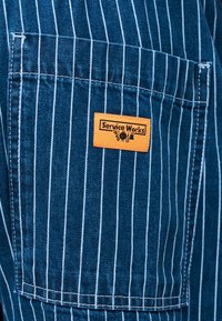 Blauwe stof met witte verticale fijne strepen en een voorkantzak met een oranje label waarop "Service Works" staat, vergezeld van kleine gereedschapsiconen.