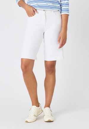 Vrouw draagt witte knielange shorts, blauw-wit gestreepte top en crèmekleurige sneakers, staat met één hand in haar zak.