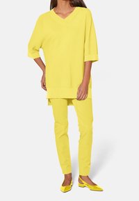 Žlutý volný svetr s výstřihem do V, žluté úzké kalhoty a žluté lakované baleríny. Hladká pletená textura, uvolněný střih, bezešvé detaily.