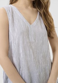 Gros plan sur une femme portant un haut sans manches blanc en V avec de fines rayures noires verticales, ses longs cheveux tombant sur son épaule.