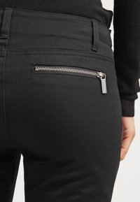 Pantalons noirs ajustés avec une texture lisse, dotés d'une poche zippée à l'arrière avec des détails en métal argenté et des coutures minimales.