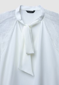 Blusa bianca con dettagli in pizzo sulle maniche corte. Presenta un design con colletto annodato e tessuto leggero e morbido. Etichetta del marchio visibile all'interno.