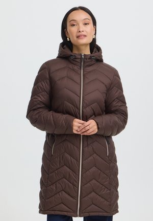 Femme portant un long manteau d'hiver matelassé marron avec capuche, fermeture éclair à l'avant et poches latérales zippées, debout les mains jointes.