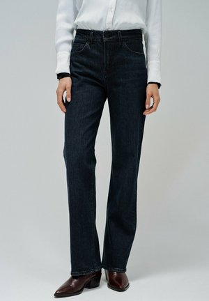 Jeans de mezclilla azul oscuro de pierna recta con cintura alta, con bolsillos frontales y detalles de costura sutiles, combinados con botas de tacón marrones.