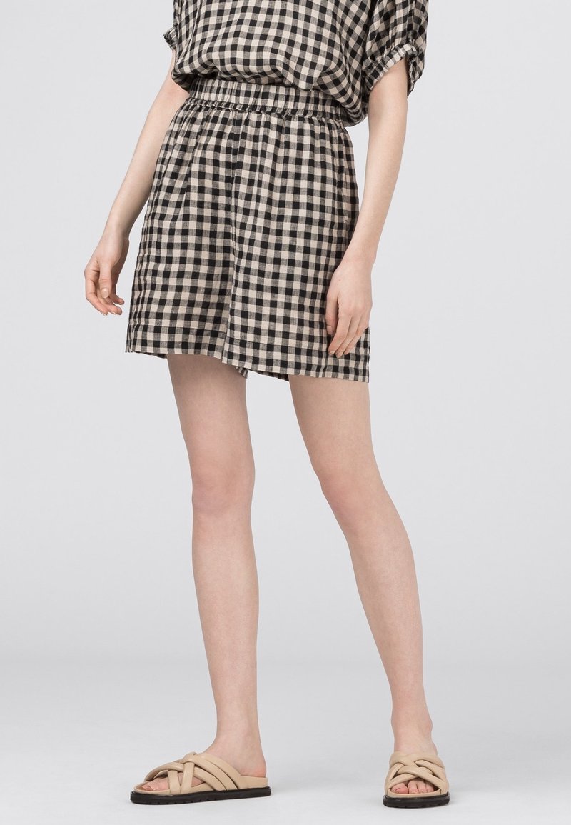 Femme portant un short et un haut à carreaux noirs et blancs avec des sandales beiges, debout devant un fond gris clair uni.