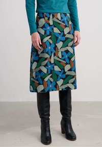 Fustă A-line multicoloră, cu un model de frunze în verde, albastru și brun pe un fundal închis; purtată cu cizme negre din piele până la genunchi.