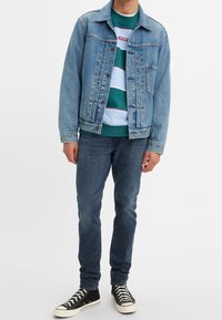 Jeansjacke in Hellblau mit verblassten Details, getragen über einem gestreiften türkisfarbenen Sweatshirt. Kombiniert mit dunklen Jeans und schwarzen High-Top-Sneakers.