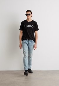 Melnas kokvilnas T-krekls ar pelēkā krāsā uzdrukātu "HUGO", gaiši zili džinsi un melnas kurpes. Modelis stāv pret neitrālu fona.