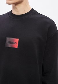 Μαύρο φούτερ από μαλακό ύφασμα, με ορθογώνιο κόκκινο και μαύρο λογότυπο με το κείμενο "HUGO" στο στήθος και ριμπ λαιμόκοψη.