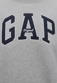 Tissu gris avec de grandes lettres bleu marine "GAP" et un mot blanc plus petit "Original" brodés au centre d'un vêtement.