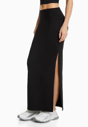Jupe maxi noire taille haute avec fentes sur les côtés, confectionnée dans un tissu doux et extensible. Portée avec des baskets blanches pour un contraste.