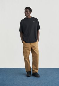 Μαύρο t-shirt με κοντά μανίκια και λογότυπο στο στήθος, συνδυασμένο με ανοιχτό καφέ παντελόνι με φαρδιά πόδια και μαύρα παπούτσια, σε ουδέτερο φόντο.