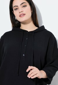 Felpa nera con texture, caratterizzata da una chiusura con bottoni, un cappuccio regolabile e maniche a tre quarti. Il design è comodo e morbido.