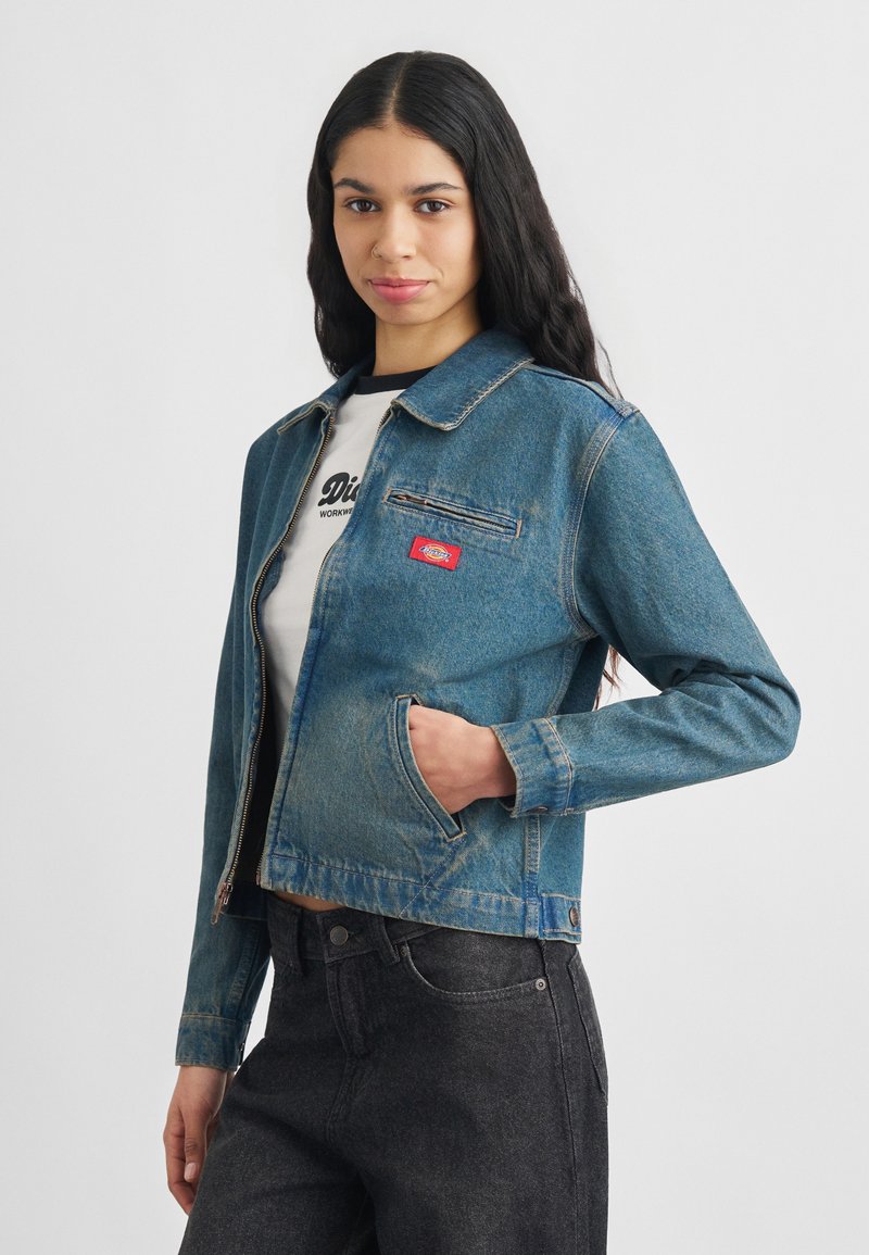 Νεαρή γυναίκα με μακριά σκούρα μαλλιά, φορώντας τζιν σακάκι σε μπλε χρώμα πάνω από λευκό t-shirt με γραφικά και σκούρα τζιν, με το χέρι στην τσέπη του σακακιού.