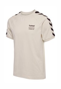 Beżowa sportowa koszulka z krótkim rękawem z czarnymi pasami w kształcie jodełki na ramionach i logo „hummel SPORTSWEAR PEANUTS” na piersi.