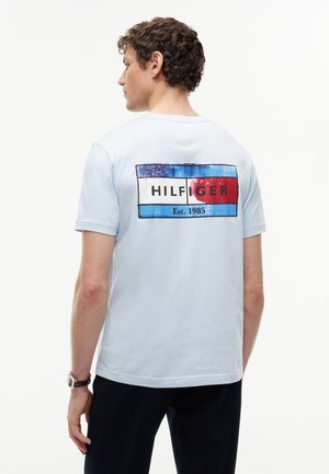 Mann mit lockigen Haaren, der ein hellblaues T-Shirt mit großem rot-weiß-blauem Tommy Hilfiger-Logo auf dem Rücken und dunkle Hose trägt.
