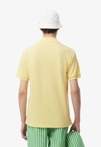 Polo di colore giallo chiaro, maniche corte, con un colletto classico. Indossato con pantaloni corti a strisce verdi e bianche e un cappello a secchiello bianco.