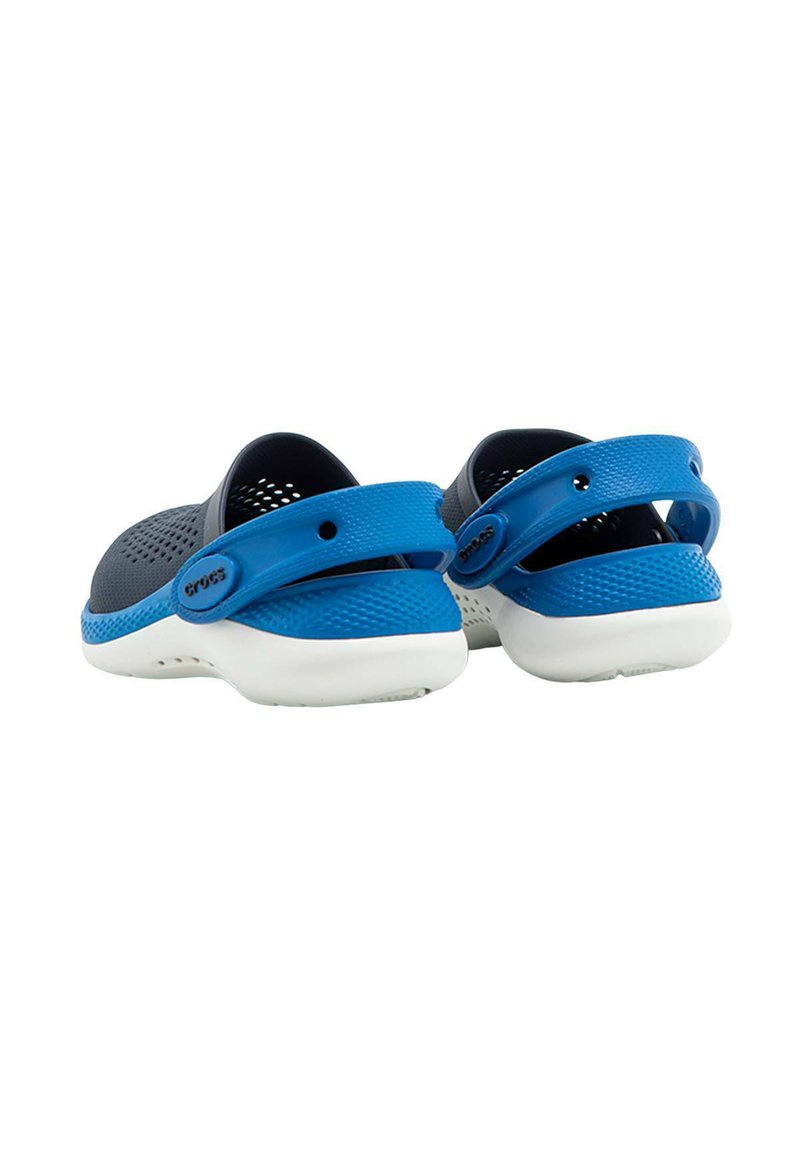 Un par de zuecos negros y azules sin cordones con correas traseras ajustables de color azul y suelas blancas, mostrados desde un ángulo trasero sobre un fondo blanco.