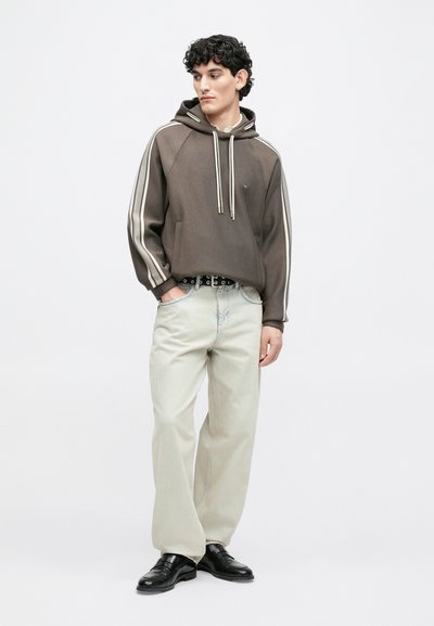 Καφέ φούτερ με κουκούλα και λευκές ρίγες στα πλάγια, σε συνδυασμό με ανοιχτό μπεζ τζιν με ίσια γραμμή και μαύρα παπούτσια, συνοδευόμενο από μαύρη ζώνη.